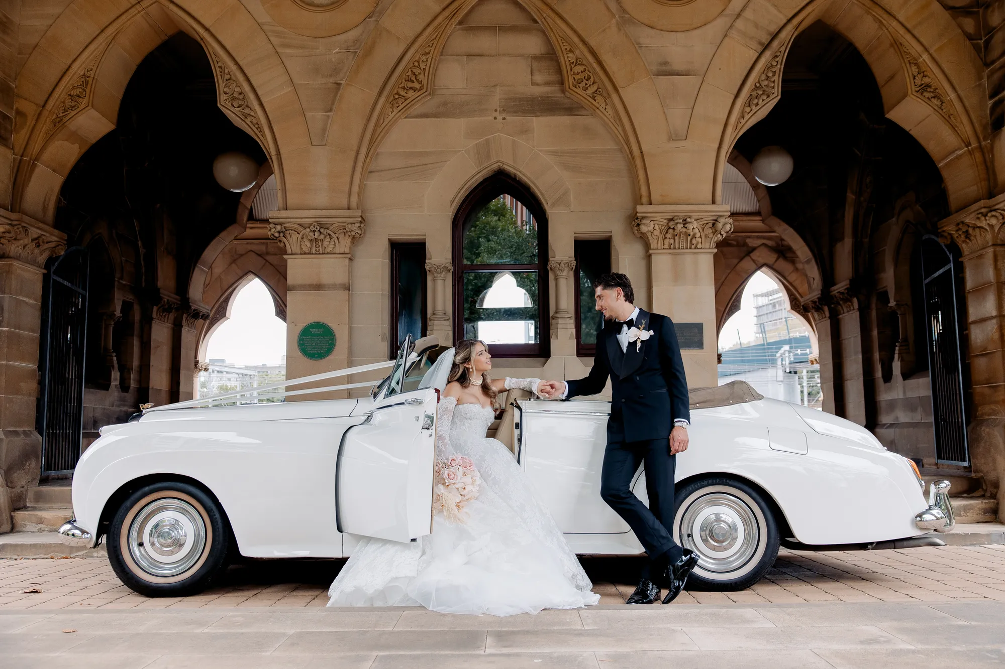 A bride and groom beside a vintage car outside a sandstone cathedral