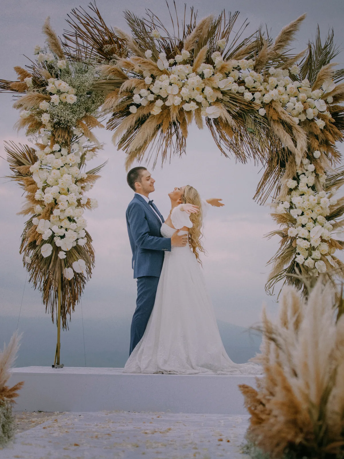 A couple beneath a pampas and white-floral ceremony arch at dusk
