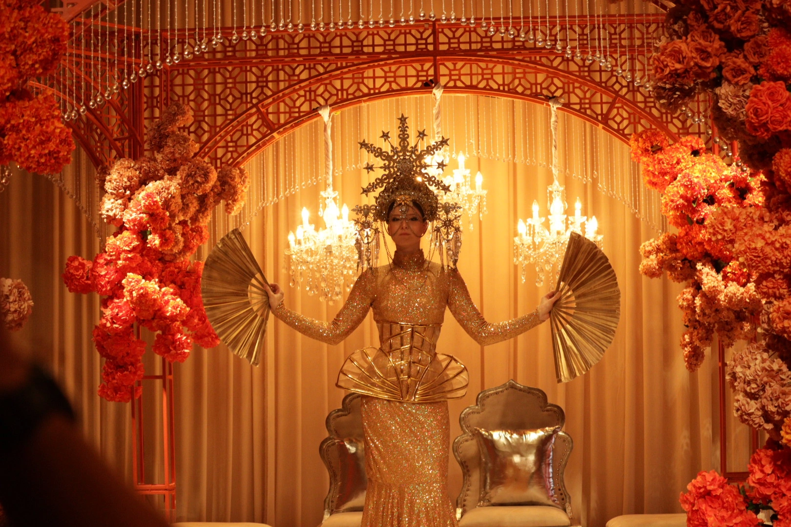 A gold-costumed performer with fans before an illuminated floral arch at Wedded World